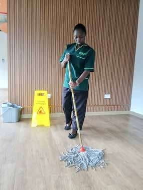 A Bitutam cleaner in branded green uniform mopping a floor beside a wet floor sign